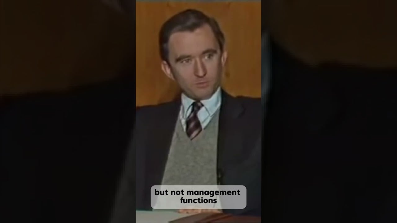 A man in a suit, vest, and tie sits at a desk with a serious expression. Subtitles read: "but not management functions." A wooden wall is visible in the background.