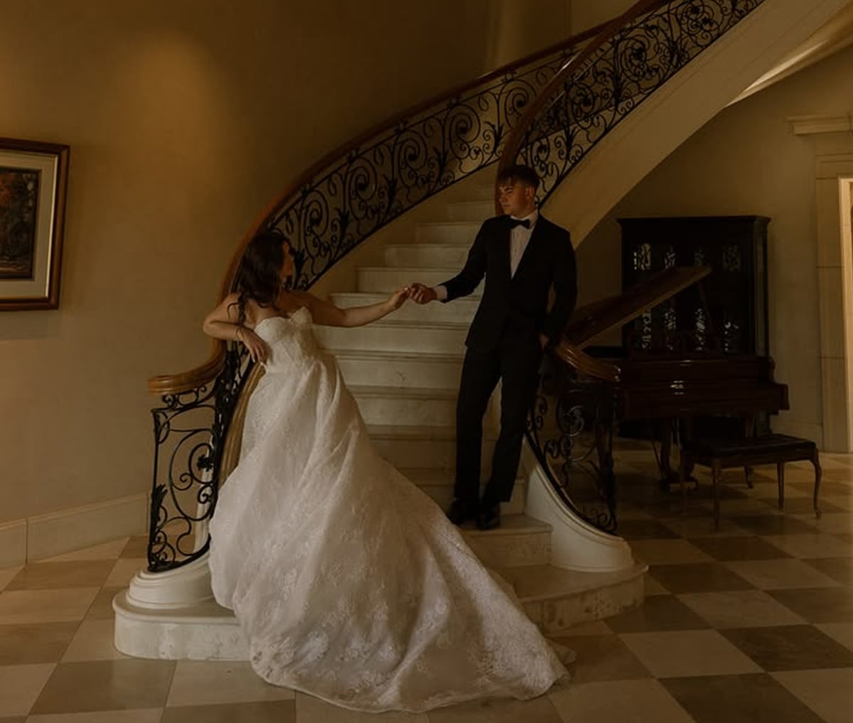 A bride in a white gown and a groom in a black tuxedo pose holding hands on a curved staircase in an elegant, warmly lit interior.