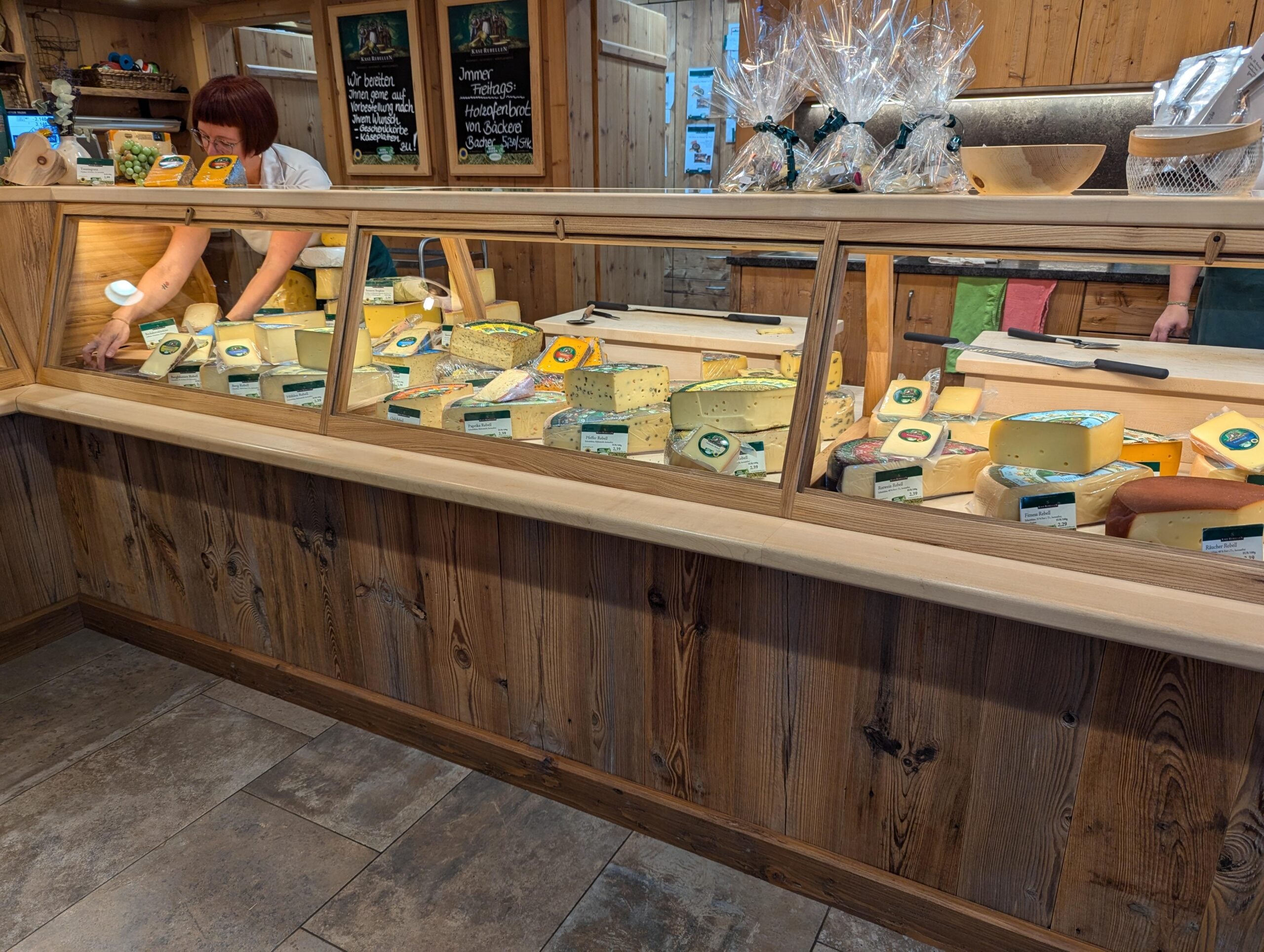 A cheese shop counter displays a variety of cheeses with labels. Behind the counter, a person slices cheese while another stands nearby. Shelves above hold wrapped cheese products, and wooden decor creates a rustic atmosphere.