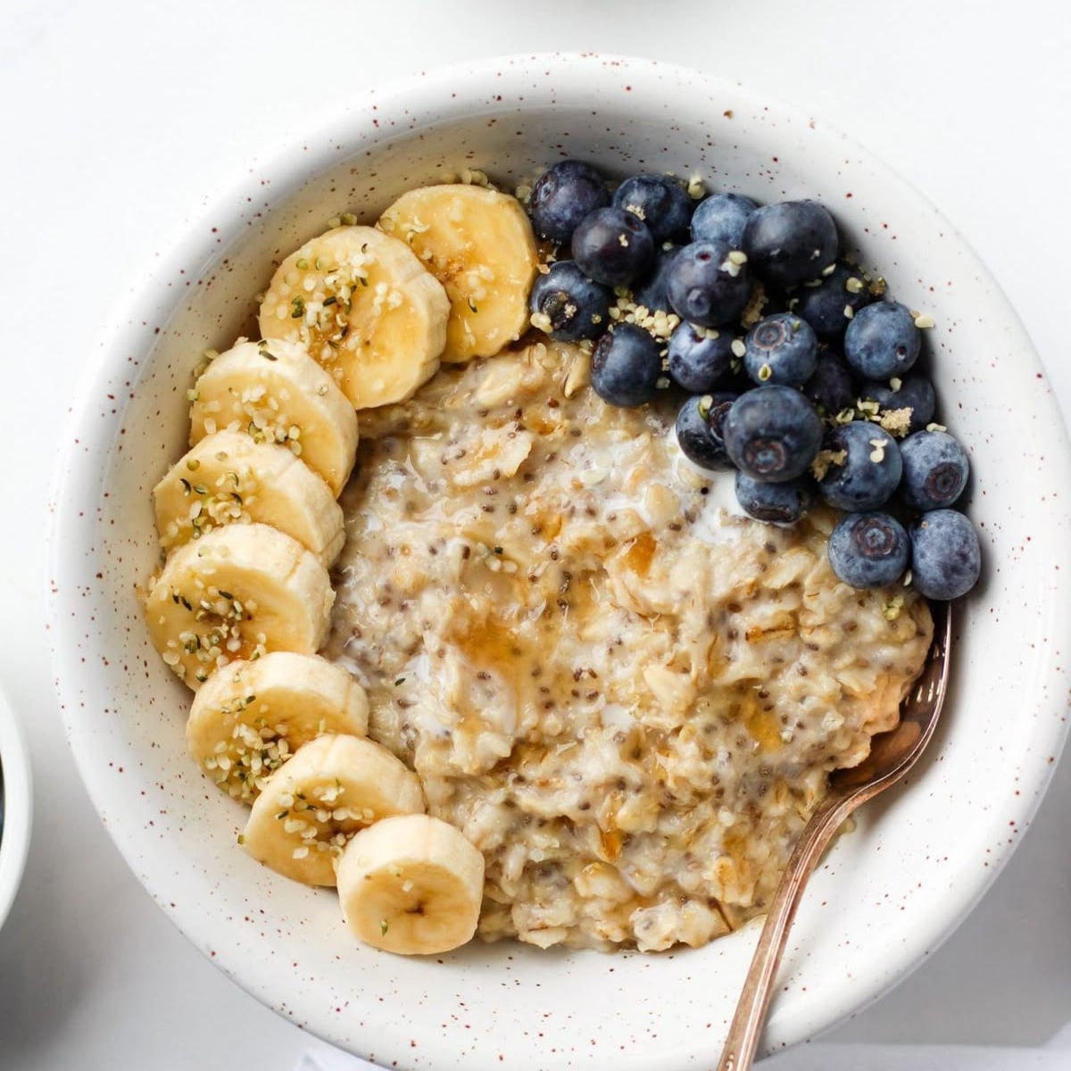 A bowl of oatmeal topped with sliced bananas, fresh blueberries, and sprinkled seeds, with a spoon resting in the bowl.