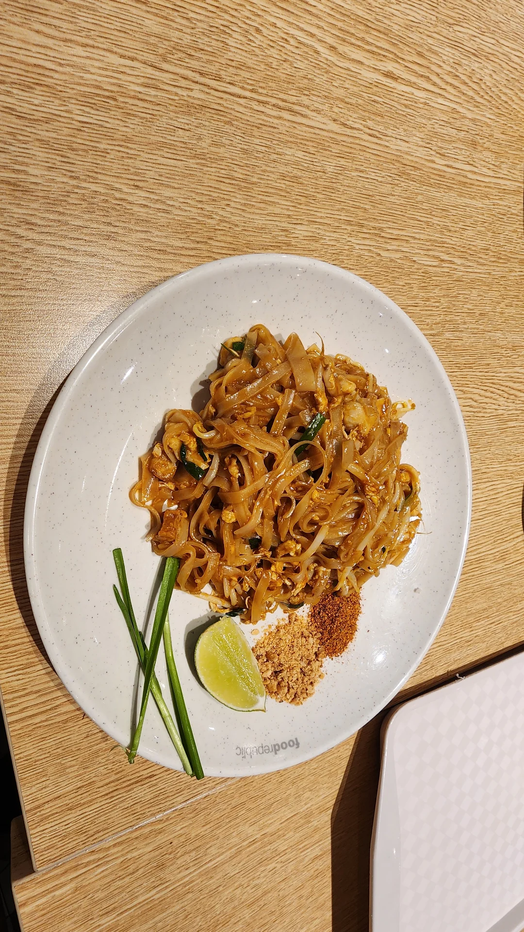 A plate of Pad Thai noodles garnished with chopped peanuts, chili powder, a lime wedge, and green onions, served on a white plate on a wooden table.