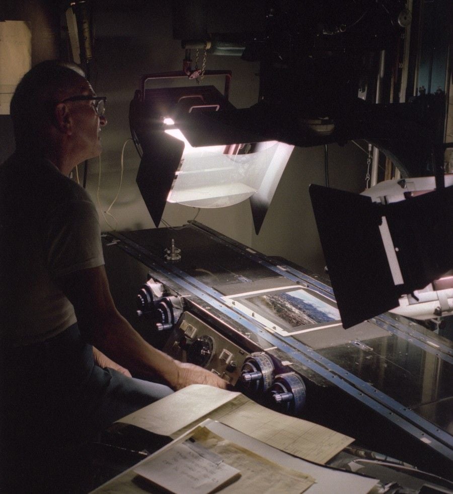 A person sits at a desk operating machinery with dials and control panels, examining documents under a large angled camera or scanner, in a dimly lit workspace.