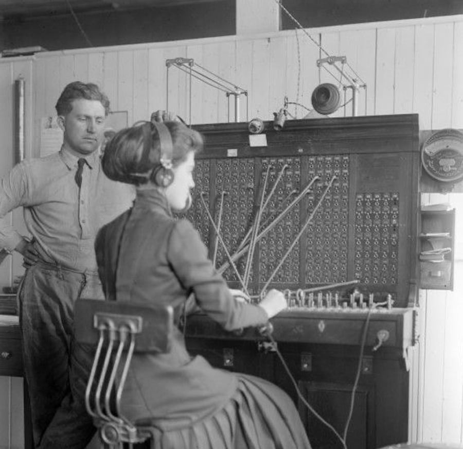 A woman operates an old-fashioned telephone switchboard, wearing a headset, while a man stands beside her watching. The setting appears to be an early 20th-century office with wood paneling.