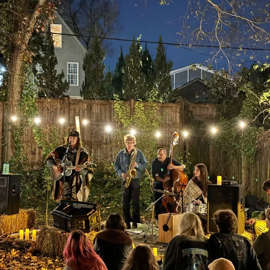 A small band performs outdoors at night under string lights, surrounded by trees and hay bales. People sit on the ground watching the musicians play guitar, saxophone, and upright bass. Candles and speakers are placed around the stage.