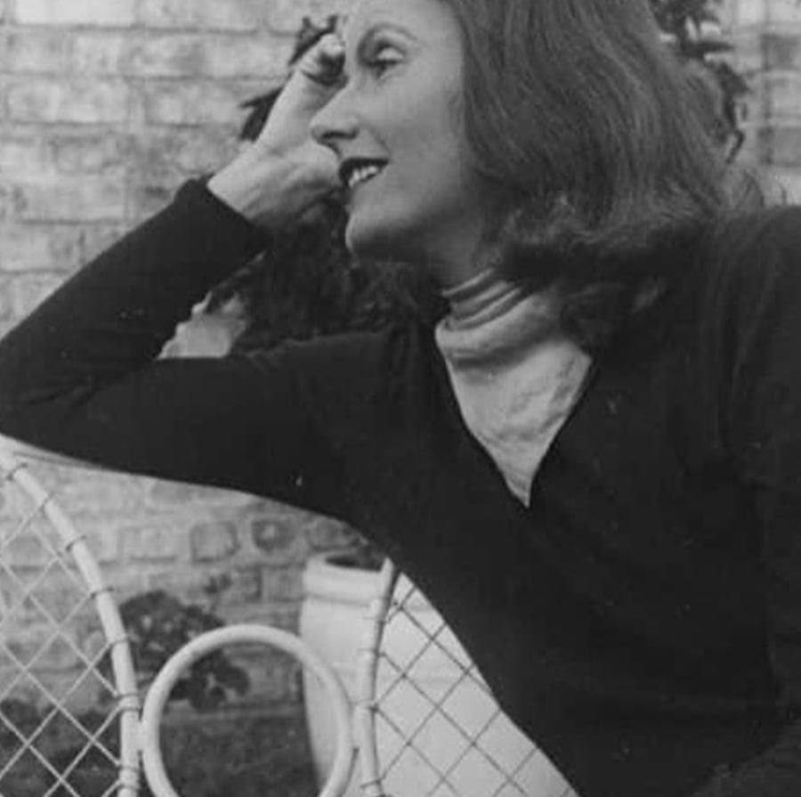 Black and white photo of a woman with long hair, sitting sideways on a chair outdoors, resting her elbow on the chair’s back and her head on her hand, smiling. There is a brick wall and a potted plant in the background.