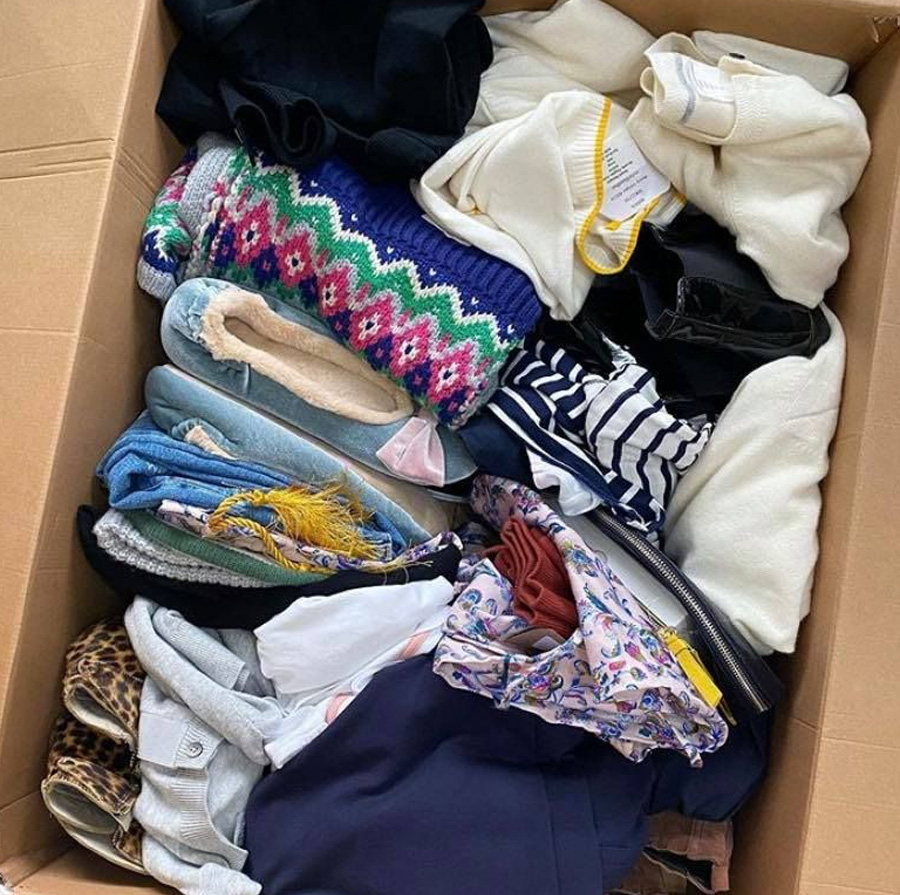 A cardboard box filled with various folded clothes, including sweaters, jeans, shirts, a striped top, and floral-patterned fabric, all neatly arranged.