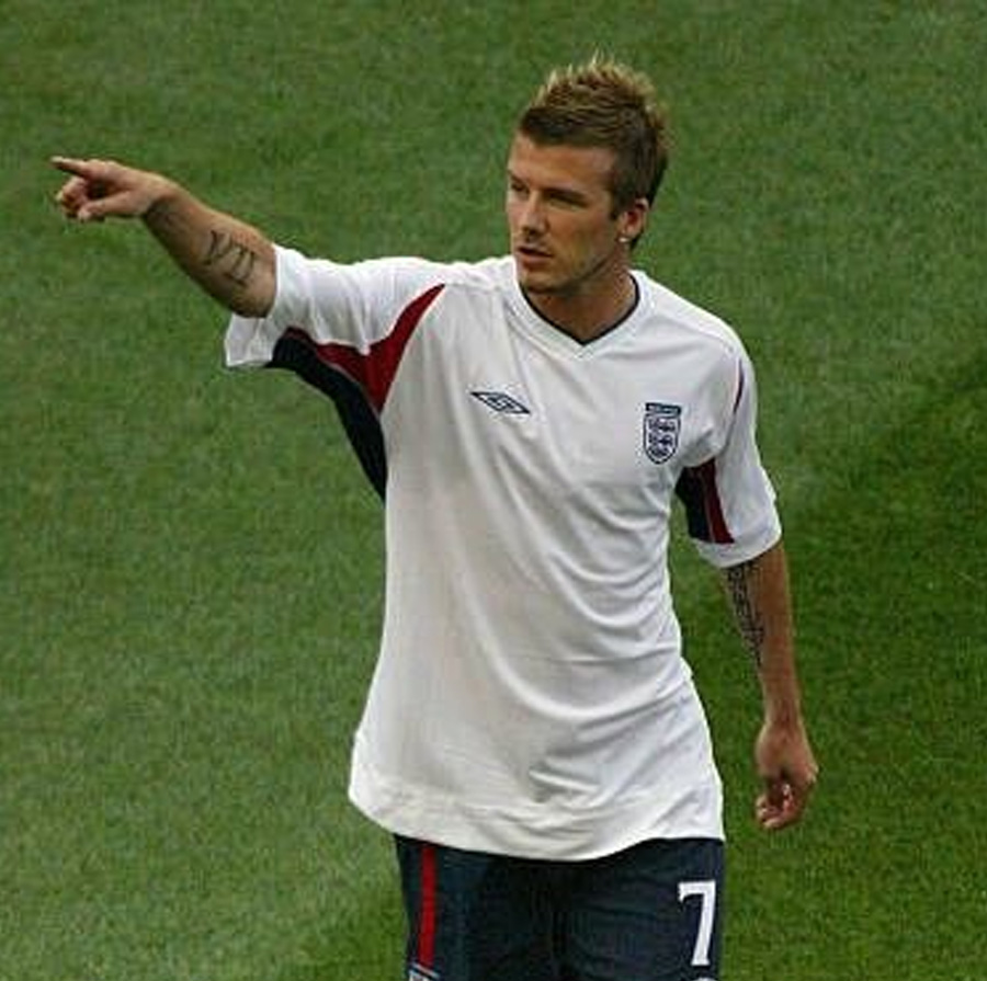 A male soccer player in an England jersey with the number 7 stands on a grassy field, pointing to his left with his right arm extended.