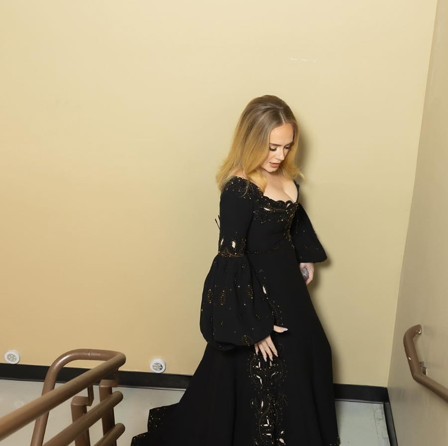 A woman in a long, off-the-shoulder black dress with gold embellishments walks down a beige stairwell, looking down and holding part of her dress.