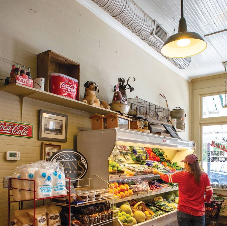 A person in a red shirt and cap selects produce from a refrigerated display in a small grocery store with vintage decor, fresh fruits and vegetables, cotton candy bags, and nostalgic Coca-Cola signs visible.