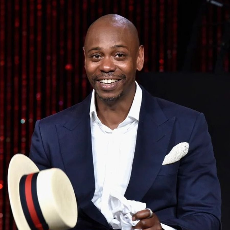 A man in a navy suit and white shirt smiles while holding a white hat with a black and red band. Red beaded curtains or lights form the background.