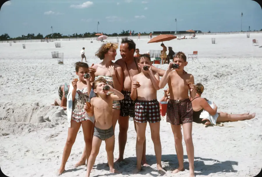 A family of six in swimsuits poses for a photo on a sandy beach. Four children in front hold cameras or toys, while two adults stand behind them. Other beachgoers and umbrellas are visible in the background.