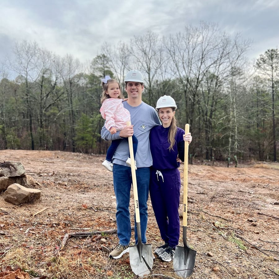 A family of three, wearing construction helmets, stands on a cleared plot of land with shovels, smiling at the camera. The man holds a young girl, and trees are visible in the background under a cloudy sky.