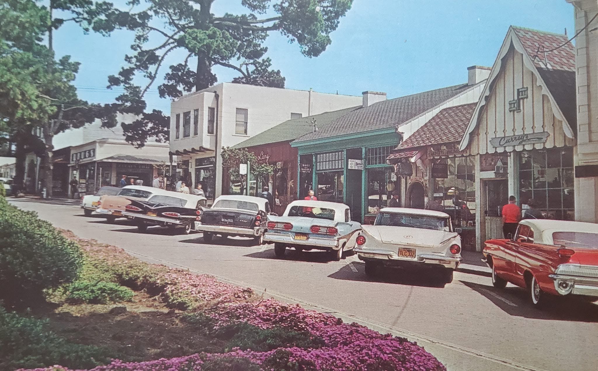 A row of vintage cars is parked along a quaint street lined with shops and cafes. Colorful flowers fill the foreground, and tall trees stand in the background under a clear blue sky.