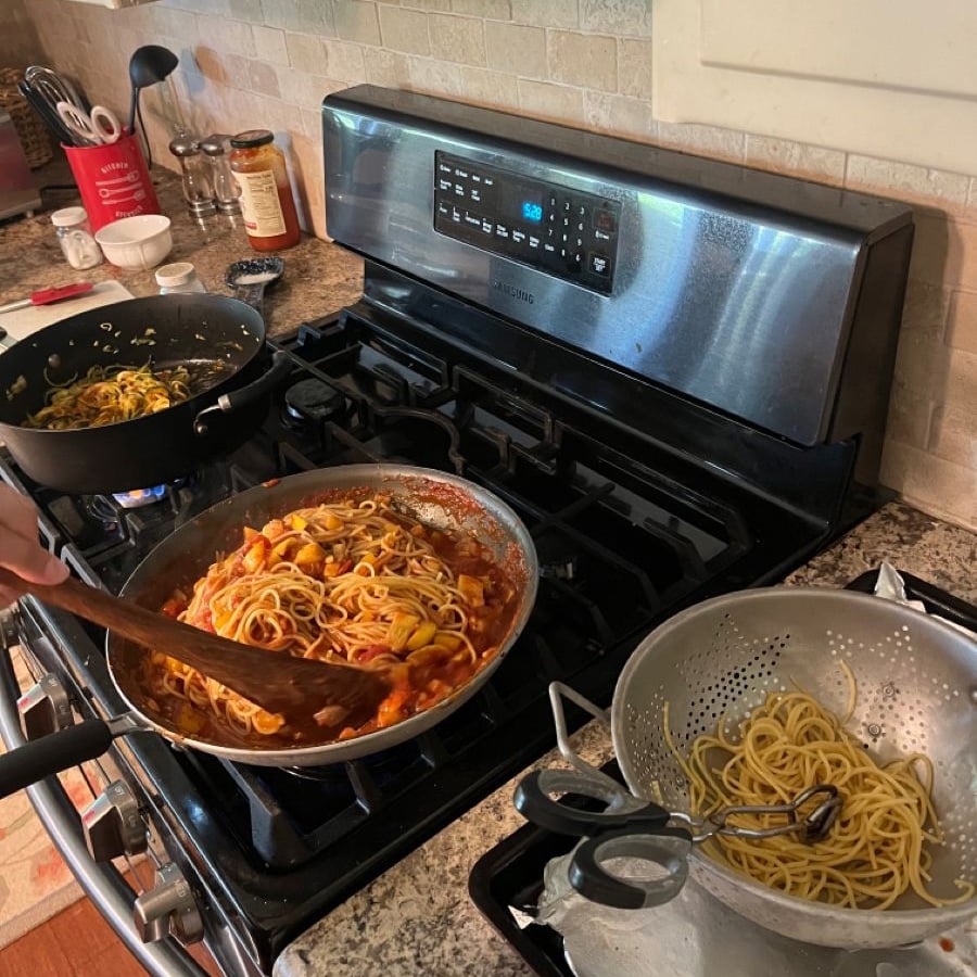 A stovetop with three pots: one with noodles and sauce, one with plain noodles, and one with noodles being stirred with a spoon. A colander with more noodles sits on the side. The stove display shows the time.