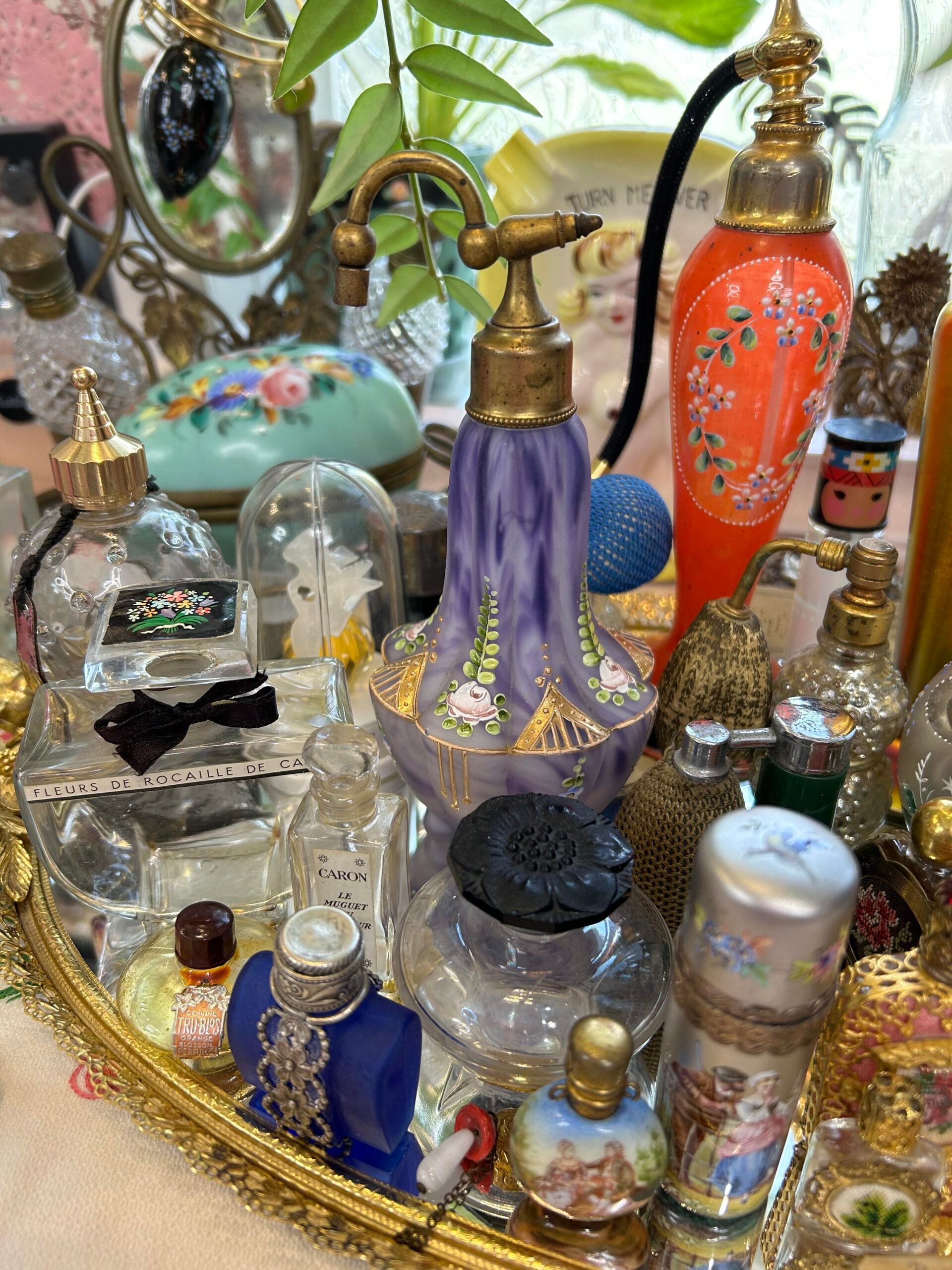 A close-up of a decorative tray filled with ornate vintage perfume bottles, many with floral designs, intricate details, and various shapes and colors, surrounded by leafy greens in the background.