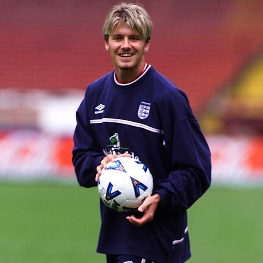 A young male soccer player in an England team training outfit smiles while holding a soccer ball on a field, with empty stadium seats blurred in the background.