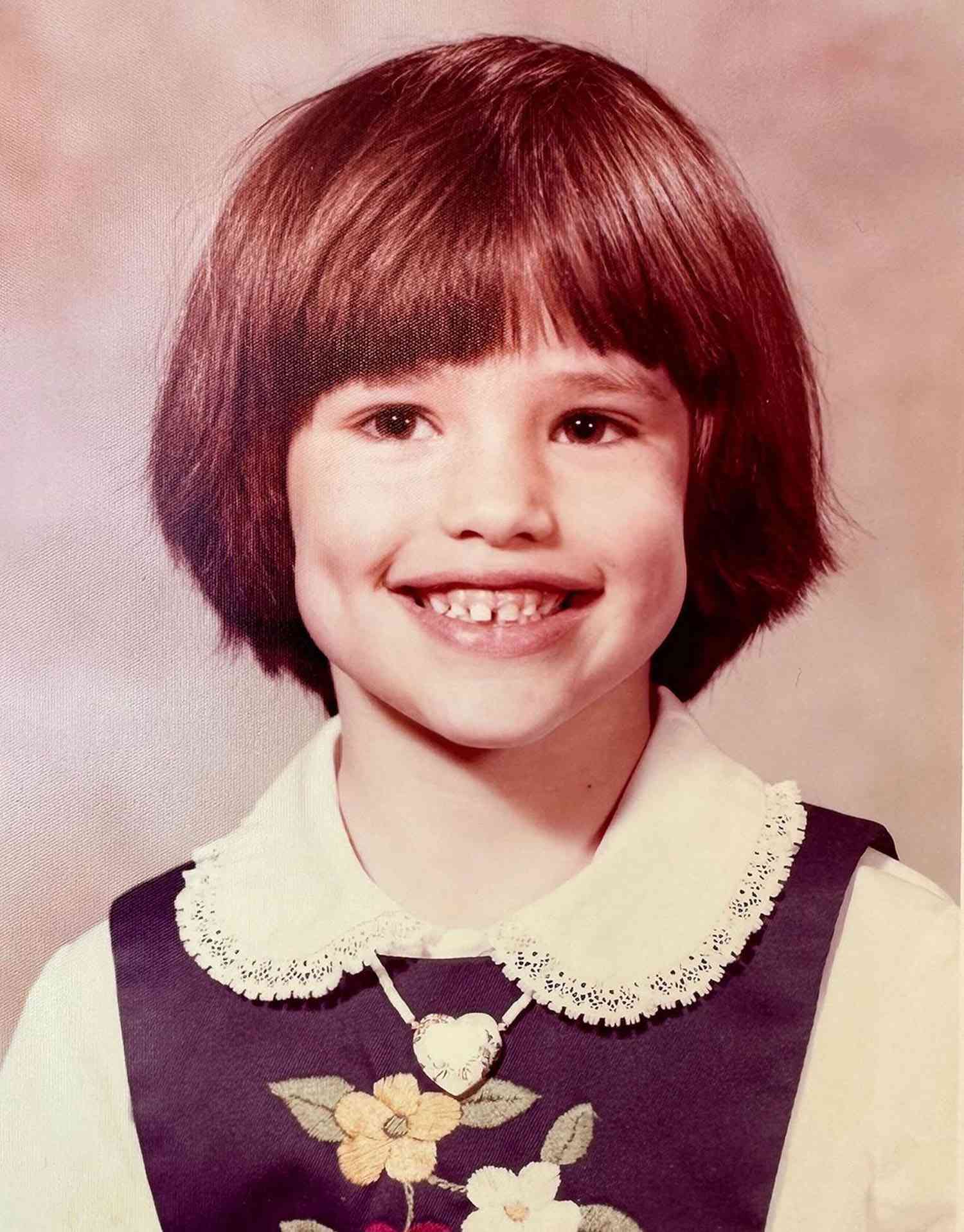 A young child with short brown hair and a wide smile shows dimples, wearing a dress with a white collar, floral embroidery, and a heart-shaped necklace, against a softly blurred background.