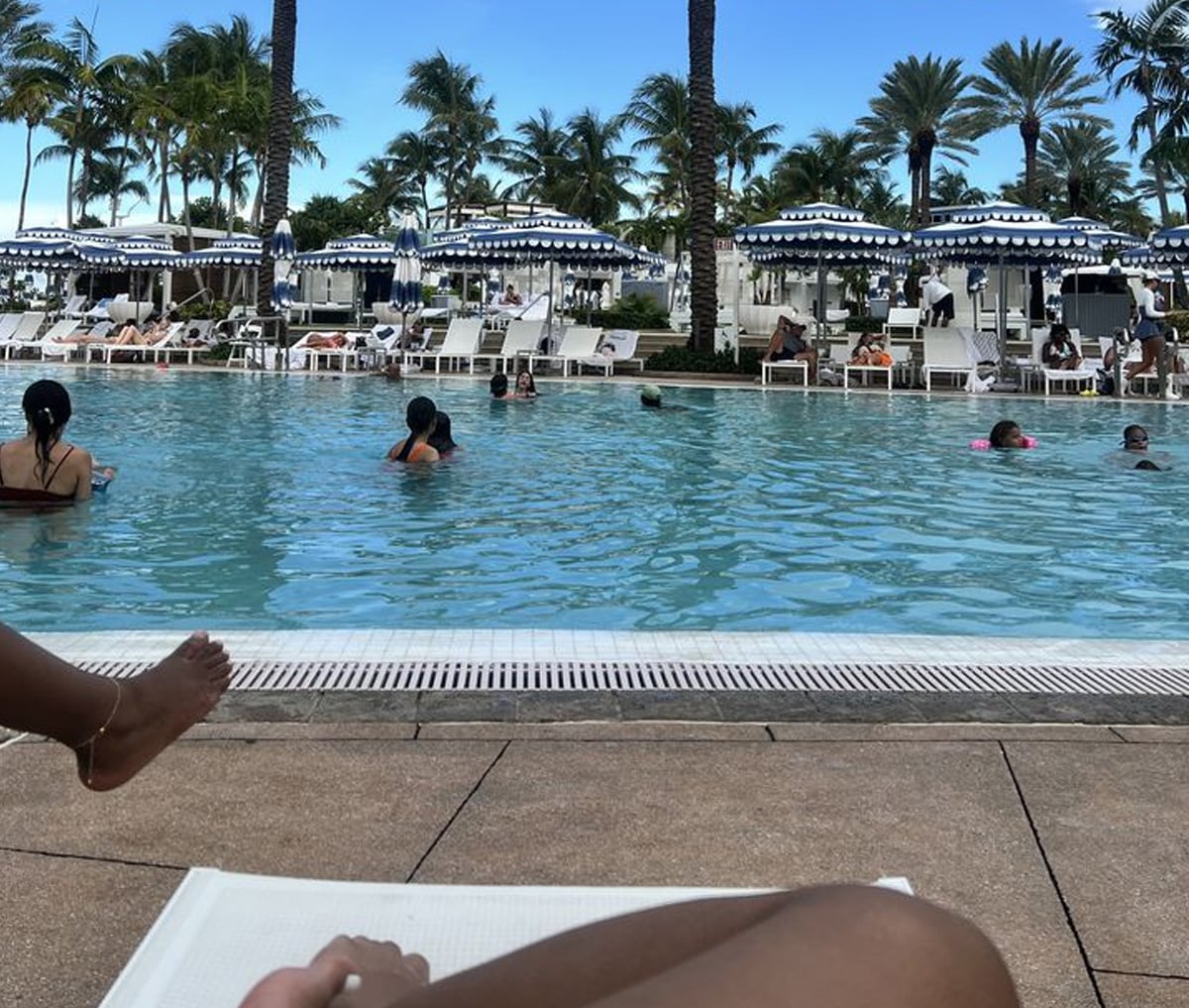 People swim and relax by a large outdoor pool surrounded by palm trees, lounge chairs, and blue-and-white striped umbrellas under a clear blue sky. Two people’s legs are visible in the foreground, resting on a lounge chair.