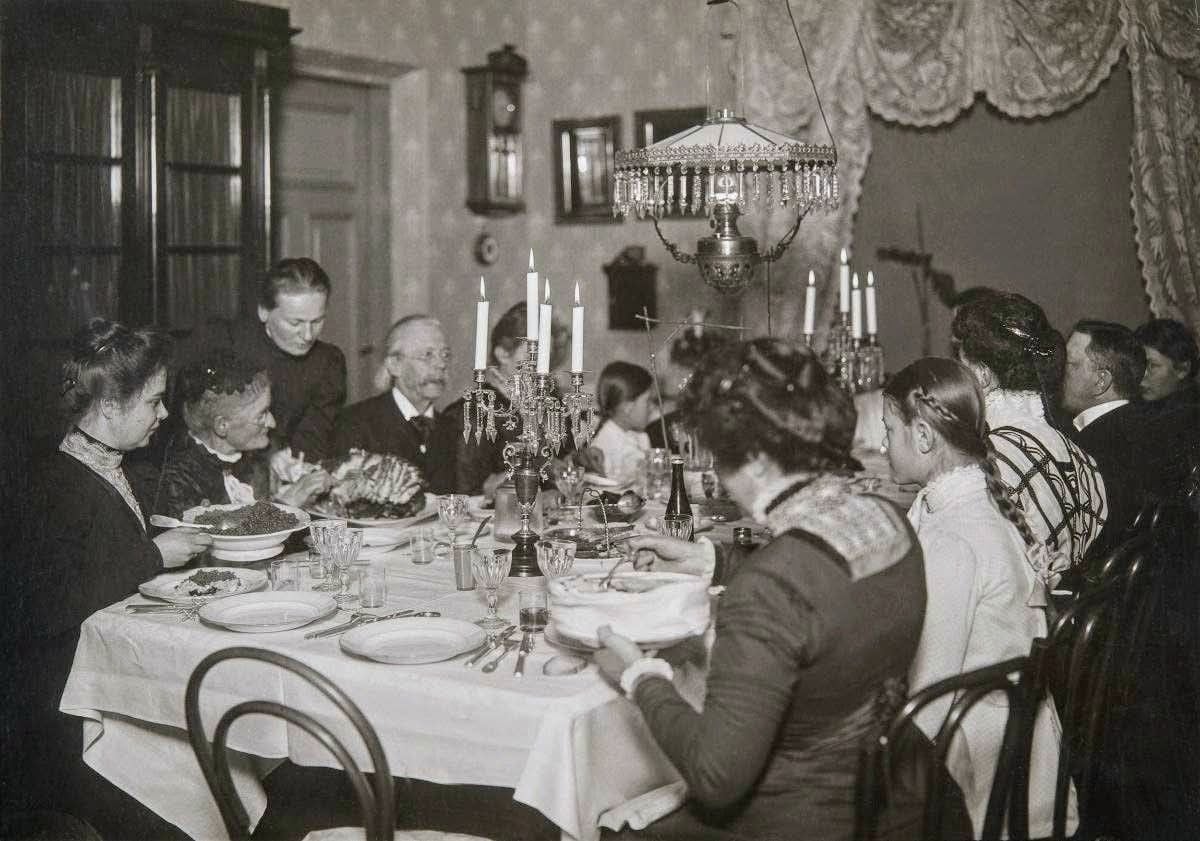 A group of people dressed in early 20th-century clothing sit around a dining table set with food, candles, and glassware in a decorated dining room with patterned wallpaper and a hanging chandelier.