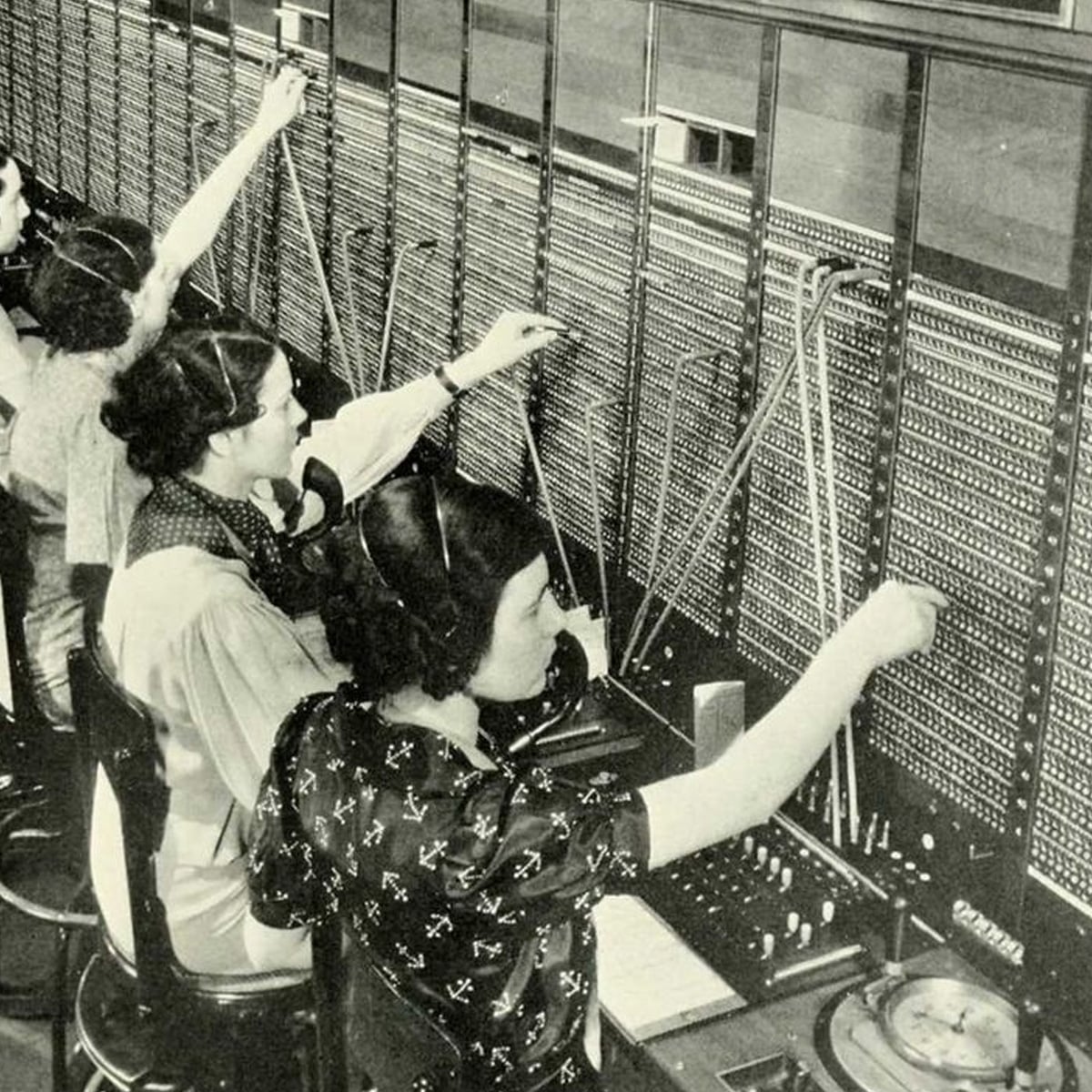 Four women wearing headsets sit in a row at a vintage telephone switchboard, connecting calls by inserting cables into various jacks on a large panel filled with wires and switches.