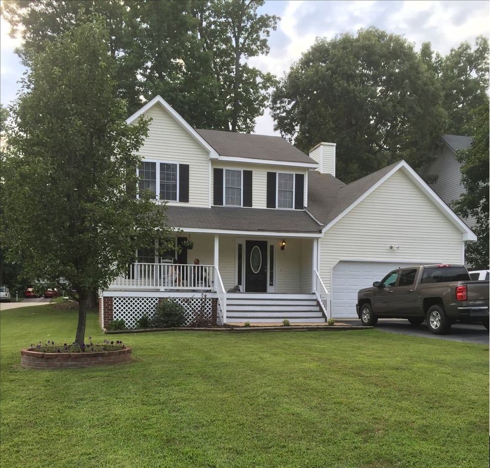 A two-story white house with a front porch, black shutters, and attached garage, sits on a well-kept lawn with a tree in front. A black pickup truck is parked in the driveway. Trees are in the background.