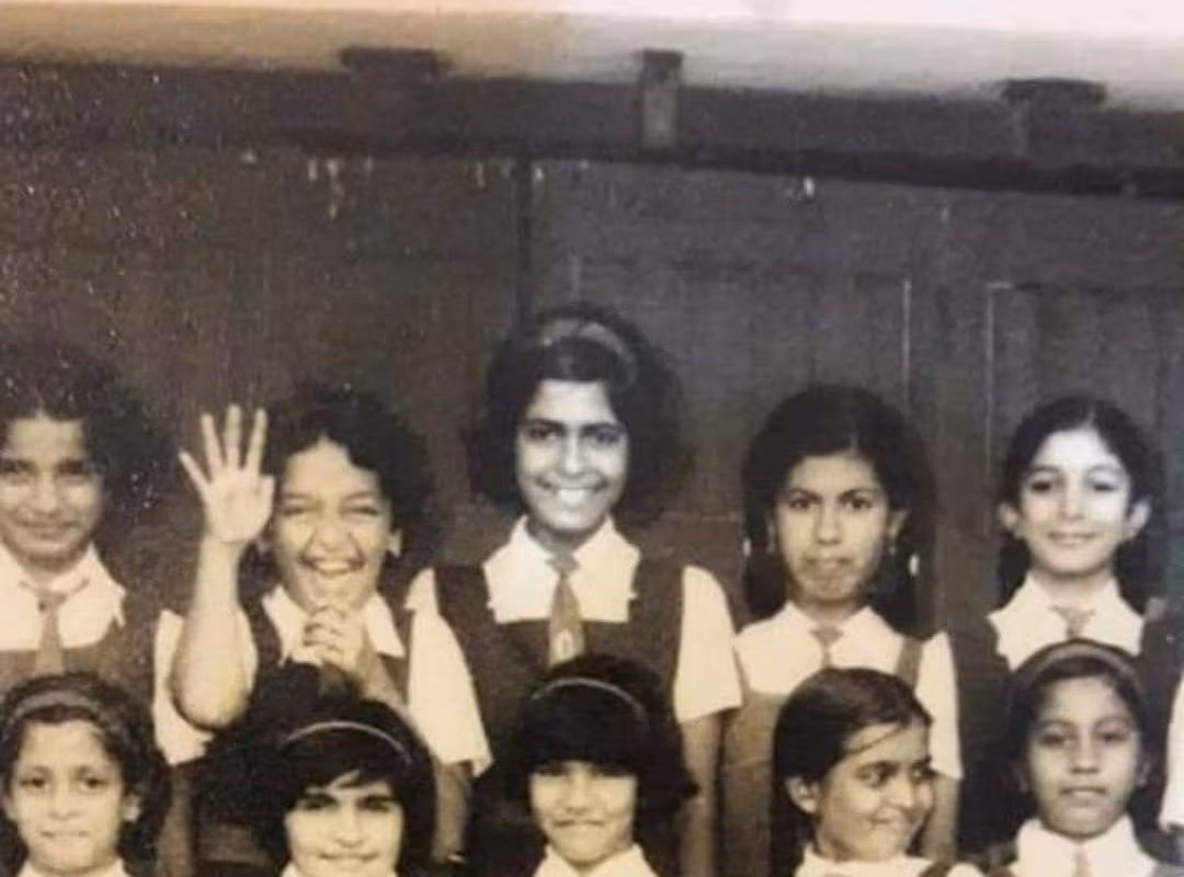 A black and white photo of ten schoolgirls in uniforms, standing and smiling for a class picture. One girl near the center is waving and several others are smiling or looking at the camera.