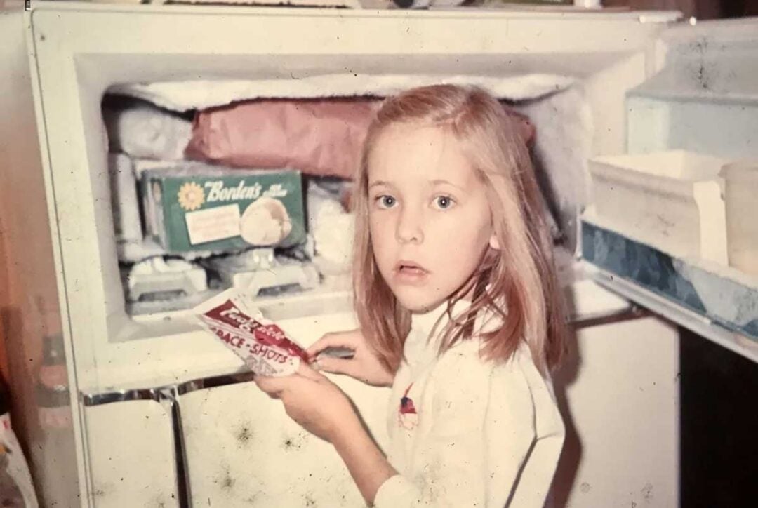 A young girl with blond hair stands in front of an open freezer, holding a package of Space Shots ice cream treats, with a surprised expression on her face.