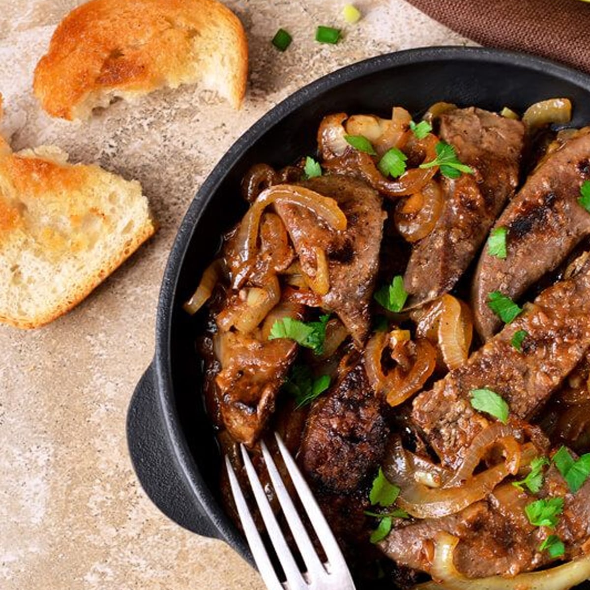 A black skillet filled with cooked liver and caramelized onions, garnished with fresh parsley. A fork rests inside the skillet. Pieces of toasted bread are placed nearby on a light-colored surface.