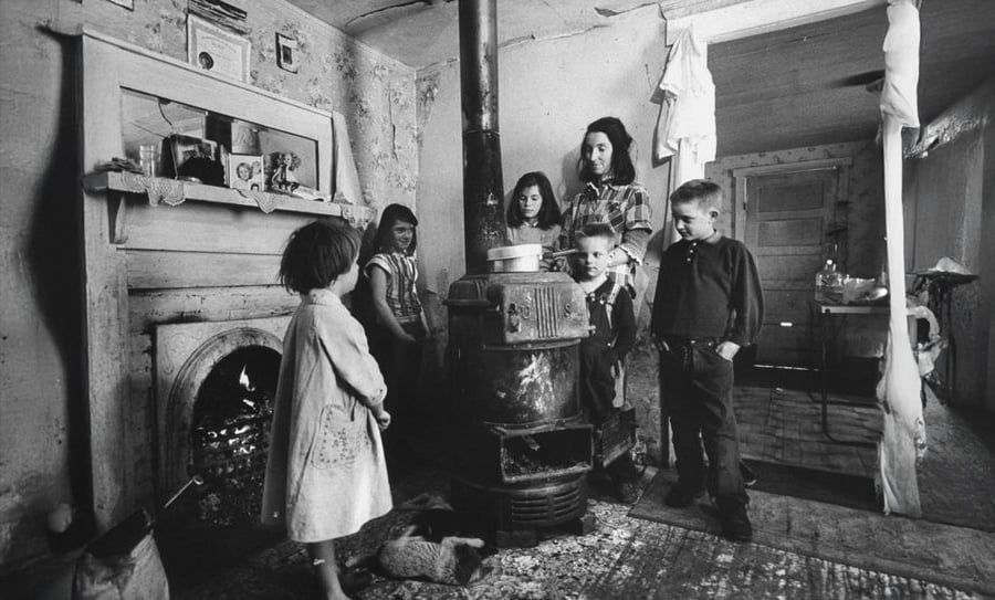 A black-and-white photo showing a woman and five children gathered around a wood-burning stove in a worn, sparsely furnished room. A cat lays on the floor nearby and a fireplace is visible on the left.