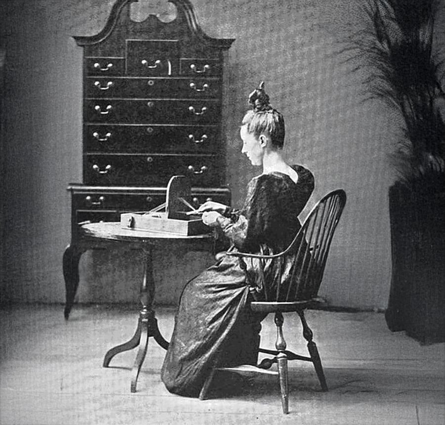 A woman in a long dress sits at a small table, writing or reading, with her hair in a bun. Behind her is a tall chest of drawers, and a large potted plant stands to her right. The setting appears vintage or historical.