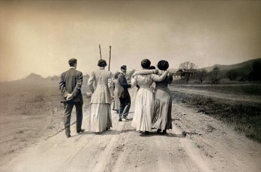 Five people in vintage clothing walk away on a dirt road. Three women walk together, two with arms around each other, and two men walk nearby. The landscape is rural, with hills and leafless trees in the background.