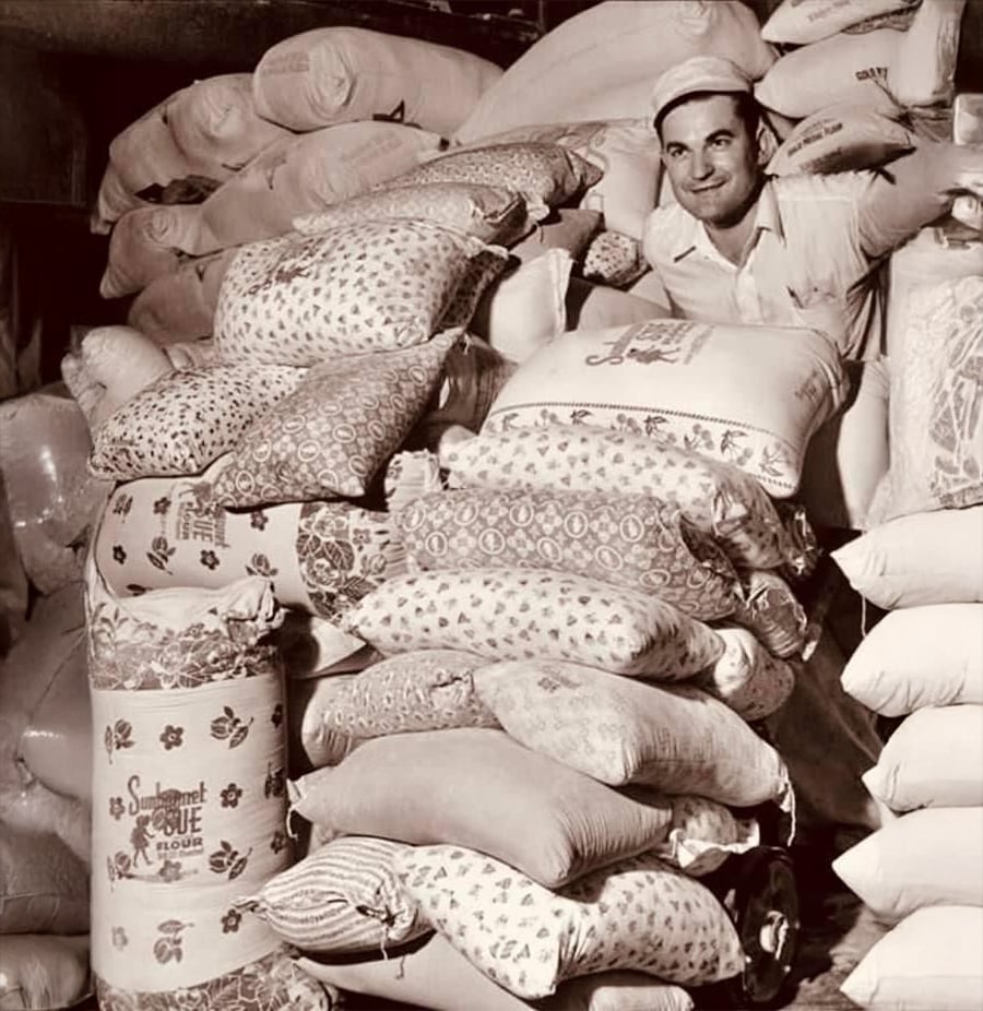 A man in a uniform and cap smiles while standing behind a large stack of patterned flour sacks in a storage area filled with more sacks. The image is in black and white.