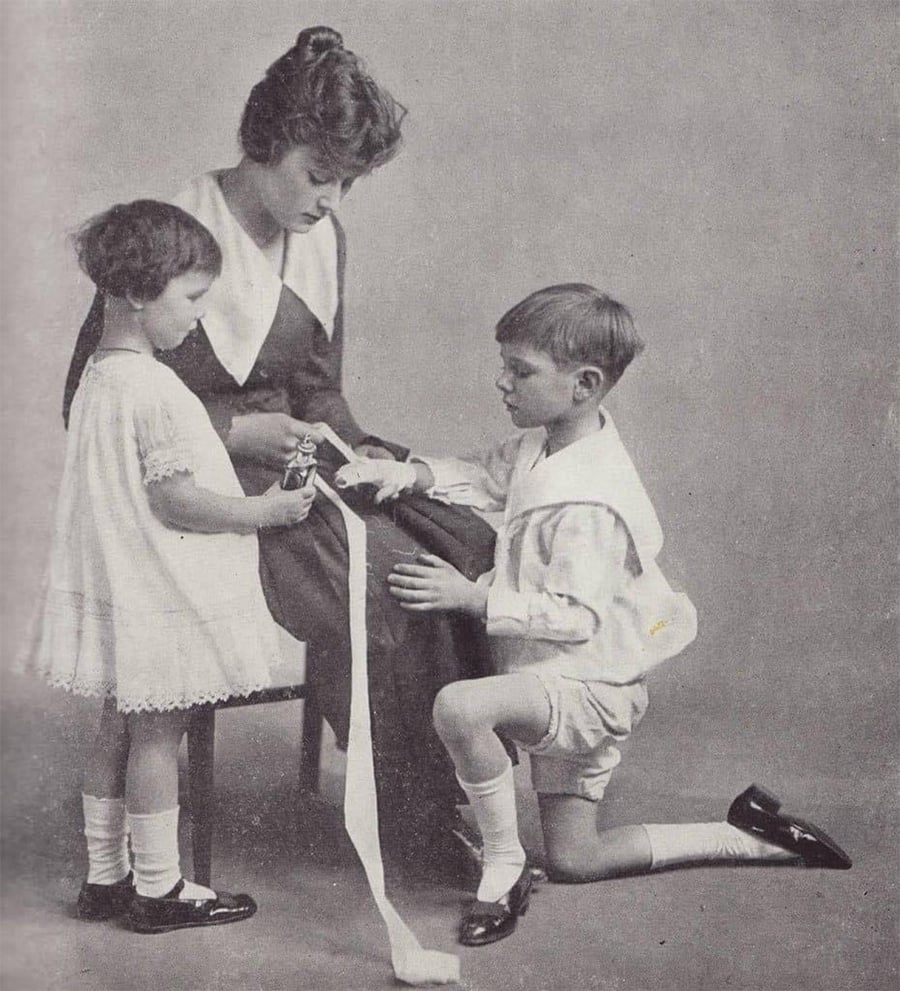 A woman sits while tending to a boy’s injured arm with a bandage. The boy kneels in front of her, and a young girl stands nearby holding a small object. All are dressed in early 20th-century clothing.