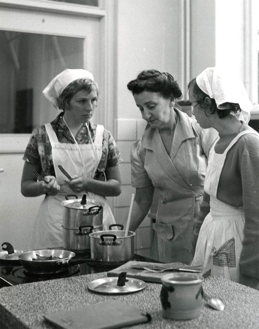 Three women in aprons and headscarves stand in a kitchen, cooking on a stove with several pots and pans. They appear to be having a discussion or cooking lesson, focused on the food.