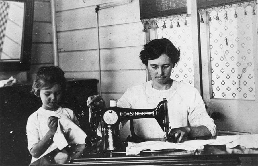 A woman uses a vintage Singer sewing machine while a young girl stands beside her, stitching fabric by hand. They are working together at a table in a room with patterned curtains.