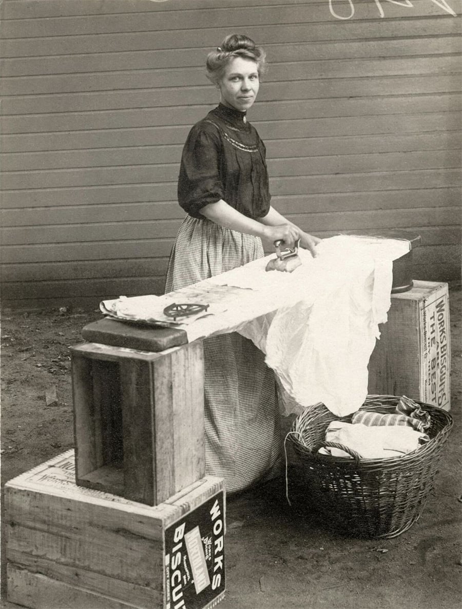 A woman stands outdoors, ironing clothes on a makeshift ironing board made of crates and a basket. She wears a long skirt and blouse, with her hair pinned up, and looks toward the camera.