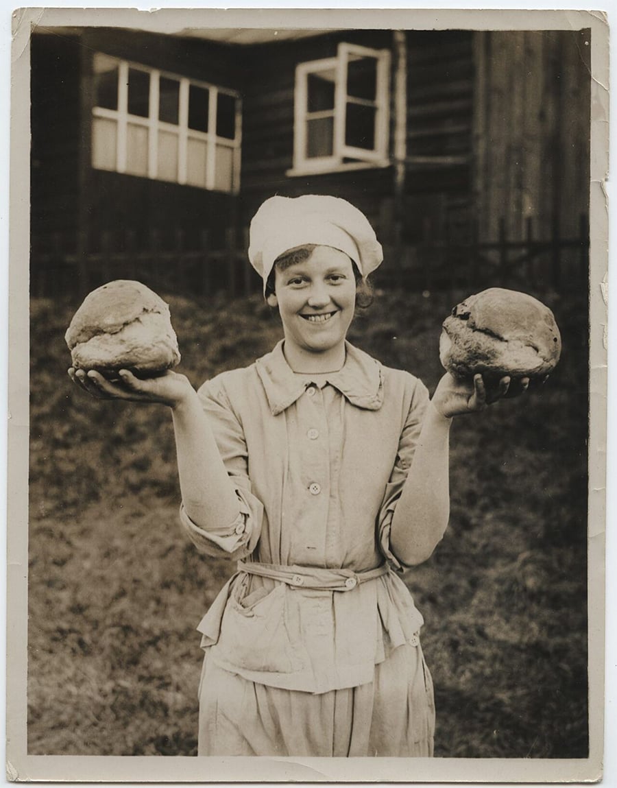 A smiling woman in a baker’s outfit and headscarf stands outdoors, holding a round loaf of bread in each hand. A wooden building with large windows is visible in the background.