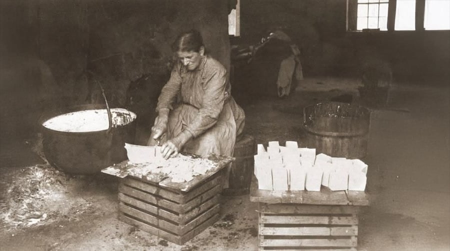 A woman in old-fashioned clothes cuts large blocks of soap on a wooden crate, with a cauldron and tub beside her and several finished bars of soap stacked nearby in a rustic, dimly lit room.