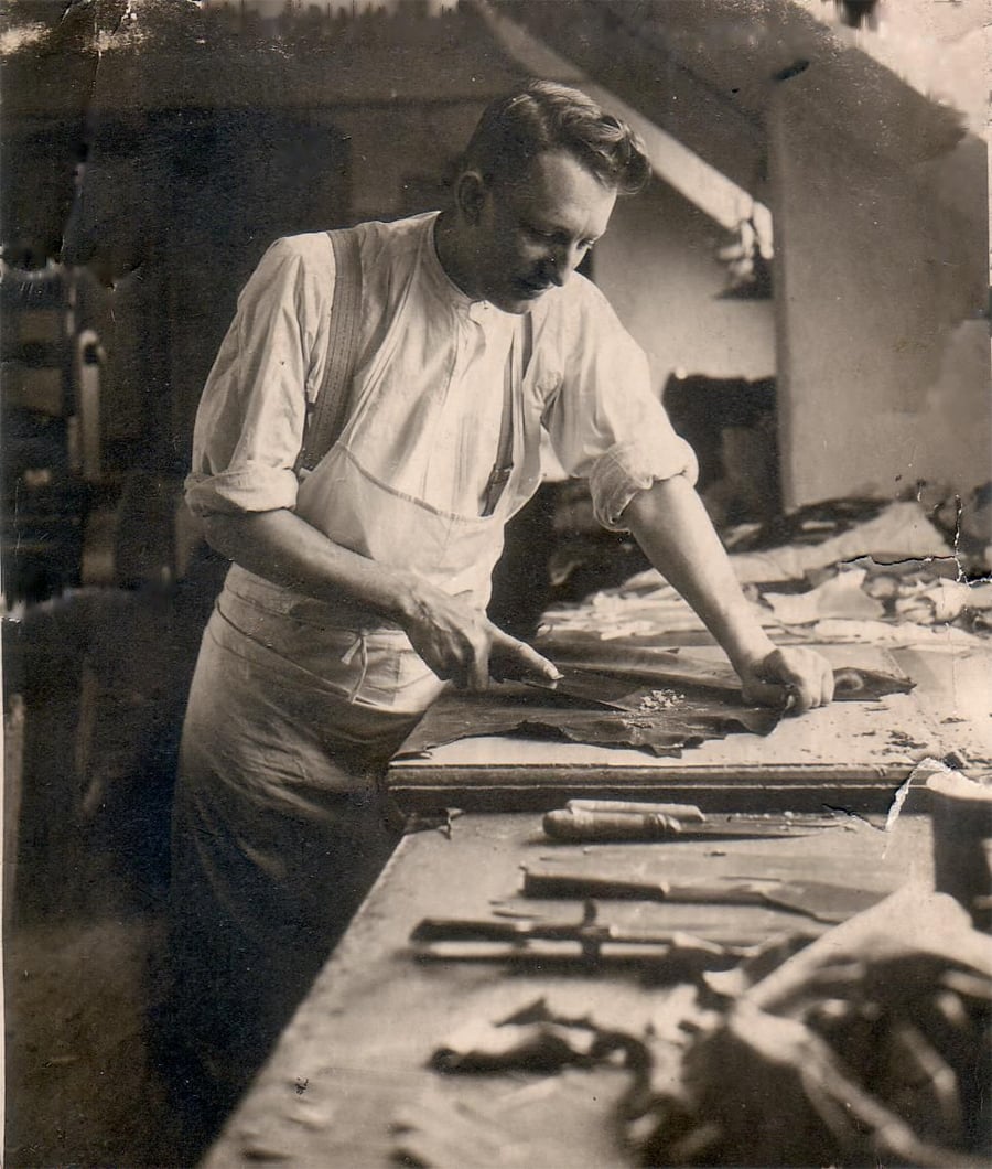 A man in a white shirt and apron works intently with a tool on a piece of material at a cluttered workbench, surrounded by various hand tools in a workshop setting.