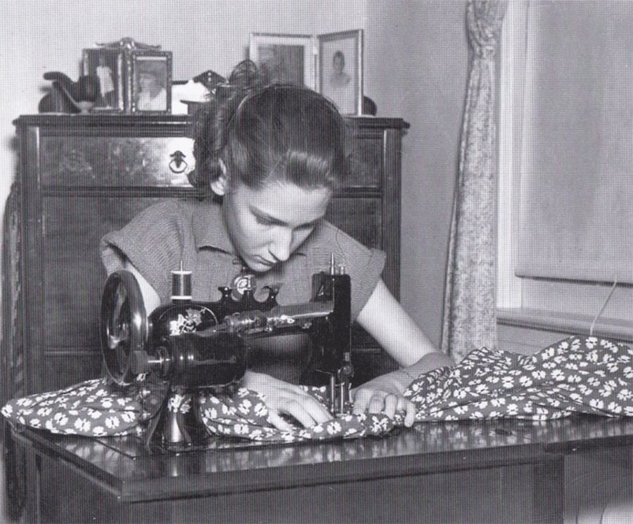 A young woman sits at a table, focused on sewing patterned fabric with a vintage sewing machine in a room with a dresser, framed photos, and a curtained window.
