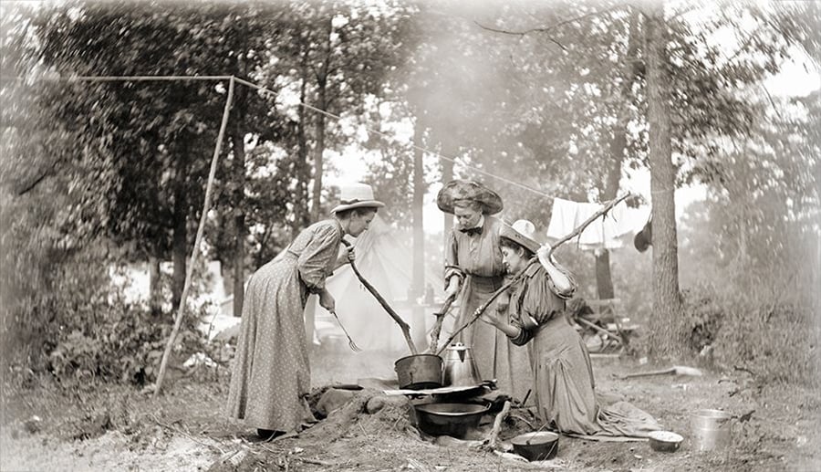 Three women in long dresses and hats cook over an open fire in a wooded outdoor setting, using large pots and utensils. The background shows trees, hanging laundry, and camping equipment. The scene appears historical.
