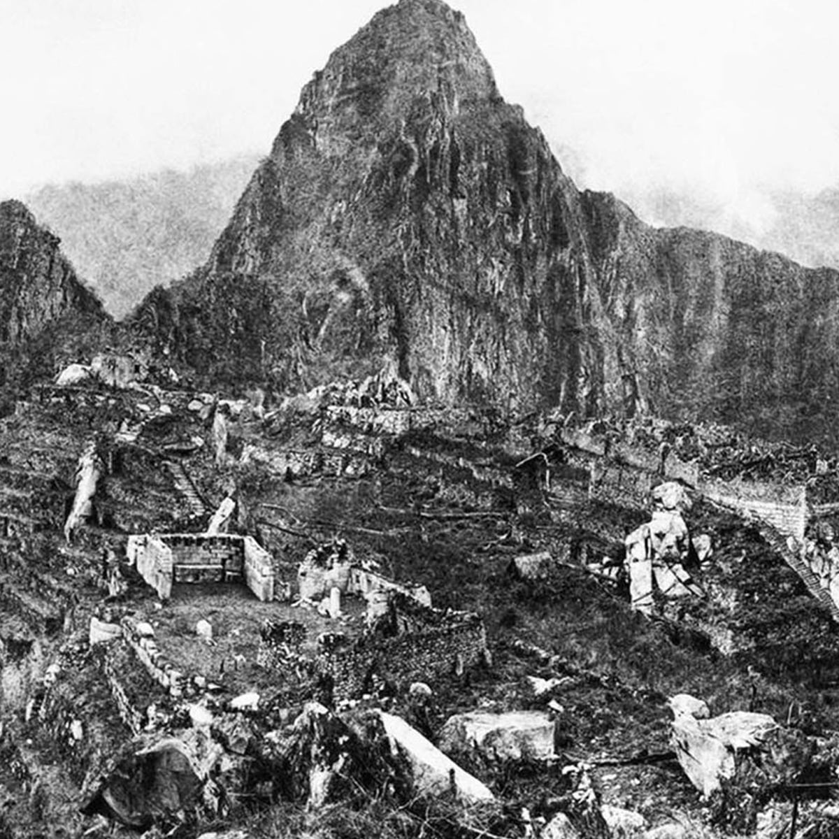 Black and white photo of ancient stone ruins set on a mountain slope with a large, steep peak rising in the background, surrounded by mist and rugged terrain.