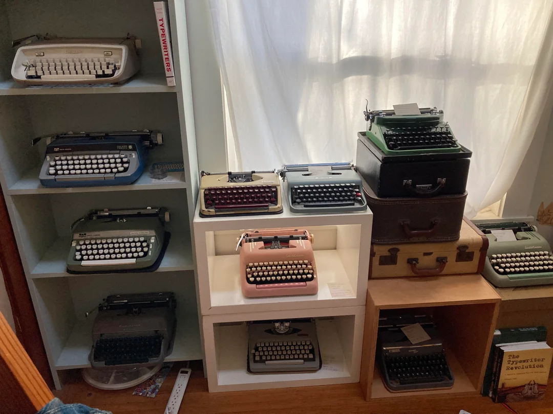 A collection of vintage typewriters in various colors is displayed on shelves and stacked on suitcases near a sunlit window, with a book titled "The Typewriter Revolution" visible on a lower shelf.
