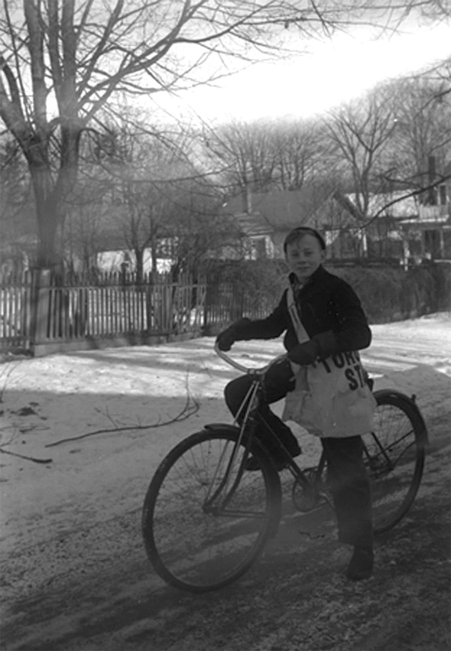 A young person stands outdoors on a snowy path, holding a bicycle. Bare trees and houses with picket fences are in the background. The person wears winter clothing and carries a bag over one shoulder.