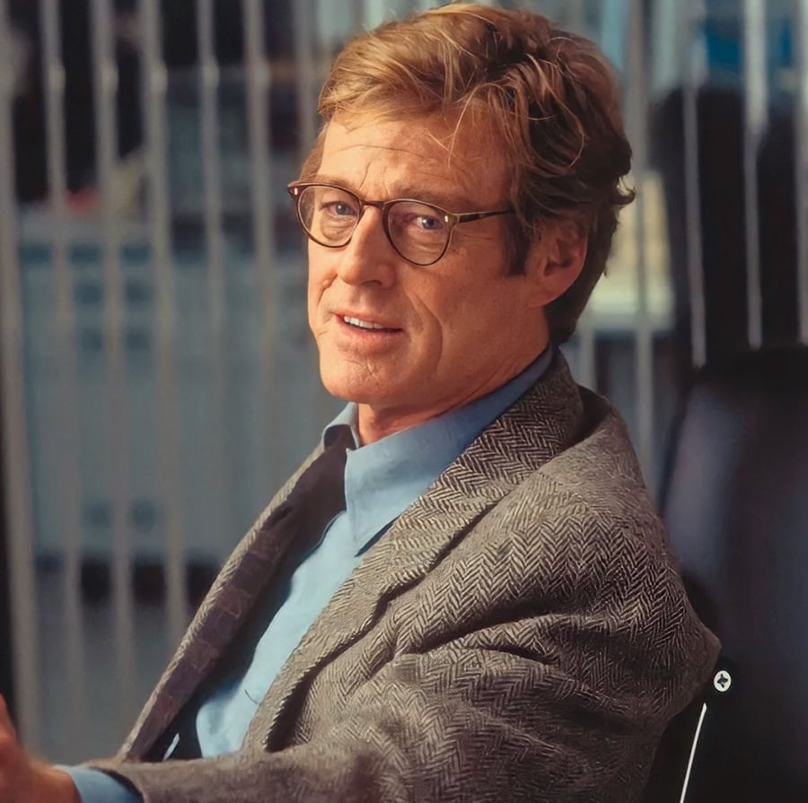 A man with light brown hair and glasses wearing a gray patterned blazer over a light blue shirt sits and looks slightly off camera, with a slight smile. Vertical blinds and an office setting are visible in the background.