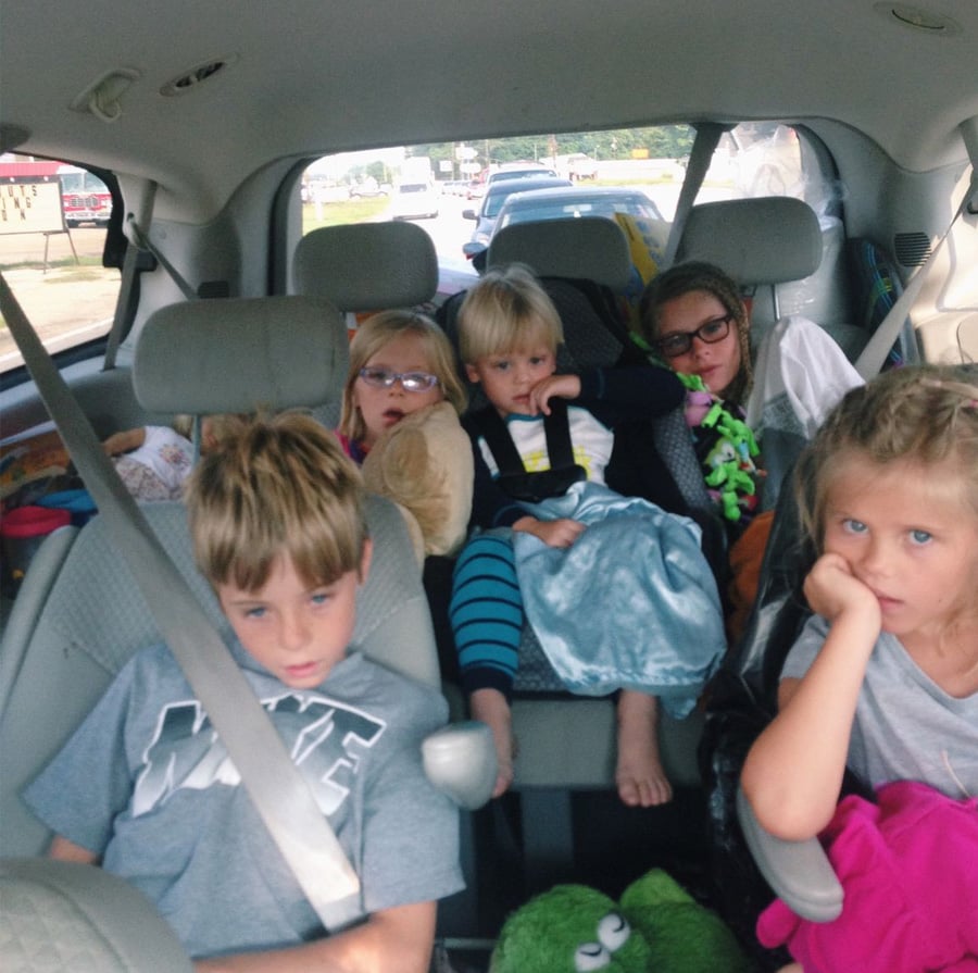 Four children sit in the back seats of a car, looking bored or tired during a road trip. They are wearing casual clothes, some with glasses and stuffed toys. Traffic and scenery are visible through the rear window.