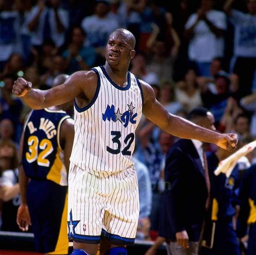 A basketball player wearing an Orlando Magic jersey with the number 32 celebrates on the court, extending his arms. The crowd and another player in a yellow uniform are visible in the background.