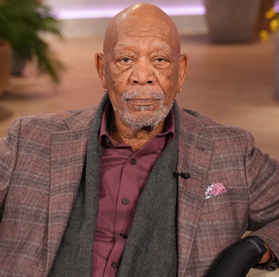 An older man with a bald head and gray beard wearing a maroon shirt, plaid jacket, and pink pocket square, sits indoors with a calm expression.