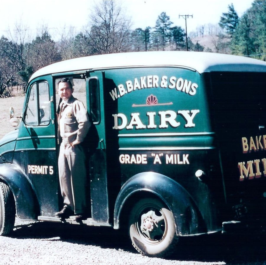A man in a uniform stands in the open doorway of a vintage W.B. Baker & Sons Dairy delivery truck marked "Grade 'A' Milk." Trees and a rural landscape are visible in the background.