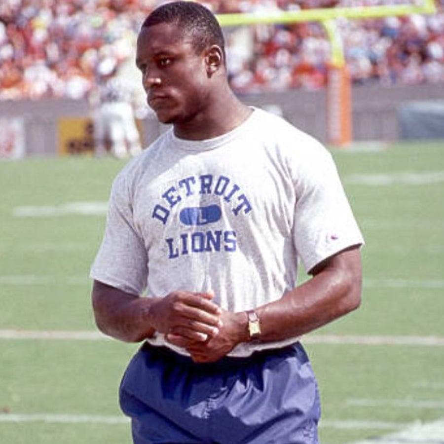 A muscular football player in a gray "Detroit Lions" t-shirt and blue shorts stands on a football field, looking thoughtfully to the side. The stadium and crowd are visible in the blurred background.